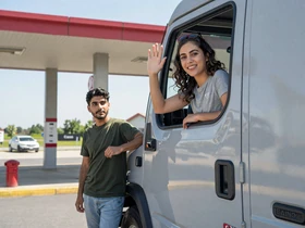 Woman waving from a truck cab window while man stands beside the truck - trucker lifestyle couple